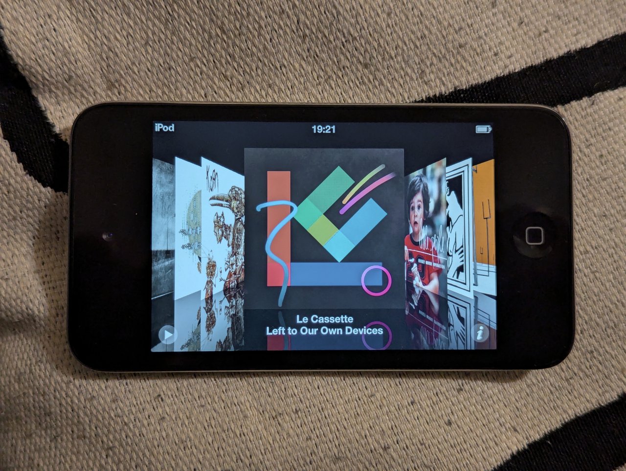 iPod in landscape mode with a gallery of album covers in cover flow. Selected is Left to Our Own Devices by Le Cassette.